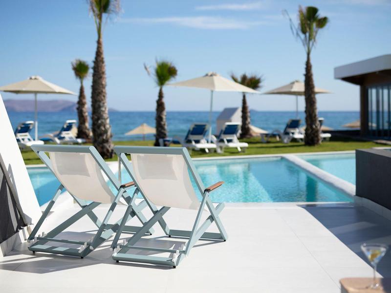 Bílé lehátka u bazénu s výhledem na moře a palmy v pozadí.