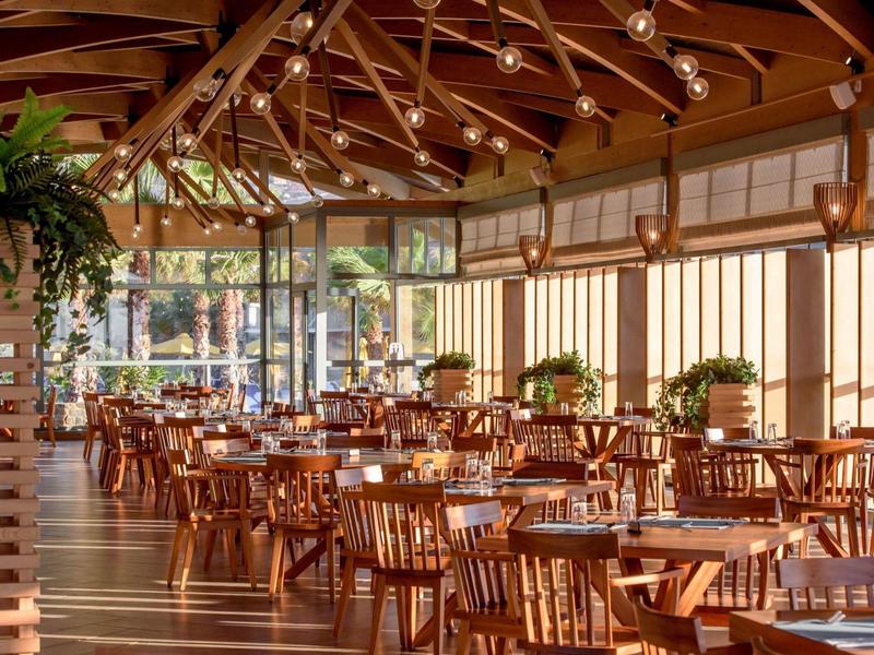 Světle osvětlená restaurace s dřevěnými stoly a velkými okny.