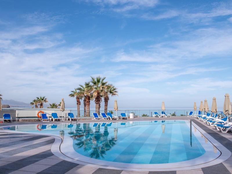 Kruhový bazén s modrými lehátky a palmami pod modrou oblohou u moře.