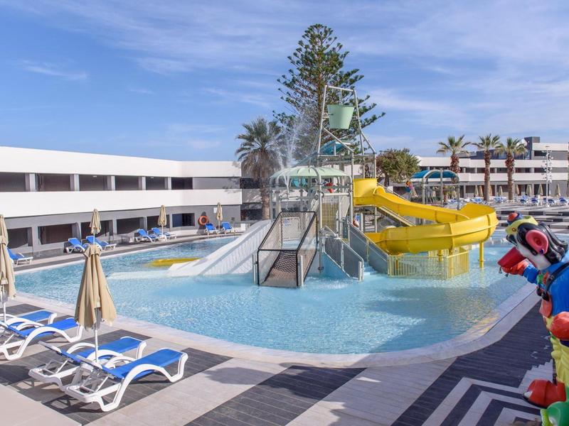 Bazén se žlutou skluzavkou a lehátky v slunném hotelovém komplexu.