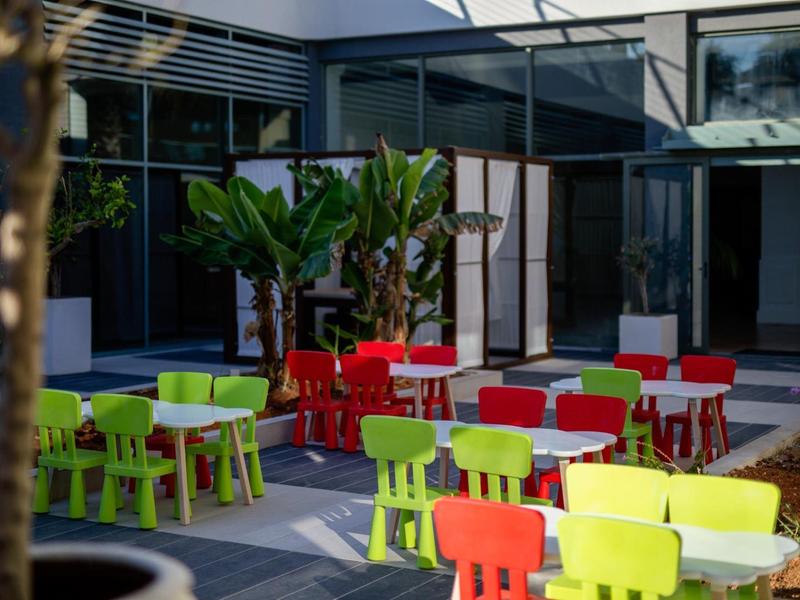 Hotelová terasa s barevnými stoly a židlemi pod velkým stromem u skleněné stěny.