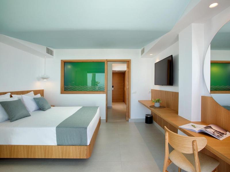 Moderní hotelový pokoj s manželskou postelí, dřevěným psacím stolem a světlým minimalistickým zařízením.