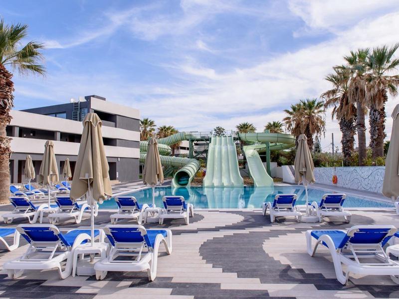 Bazén s tobogány a lehátky pod modrou oblohou, obklopený palmami.