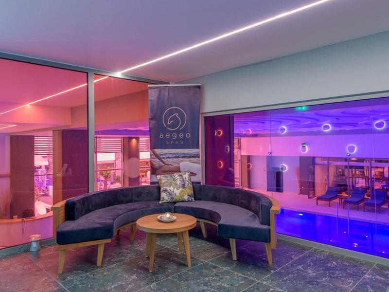 Pohodlný salón s kulatou pohovkou a výhledem na vnitřní bazén osvětlený fialově.