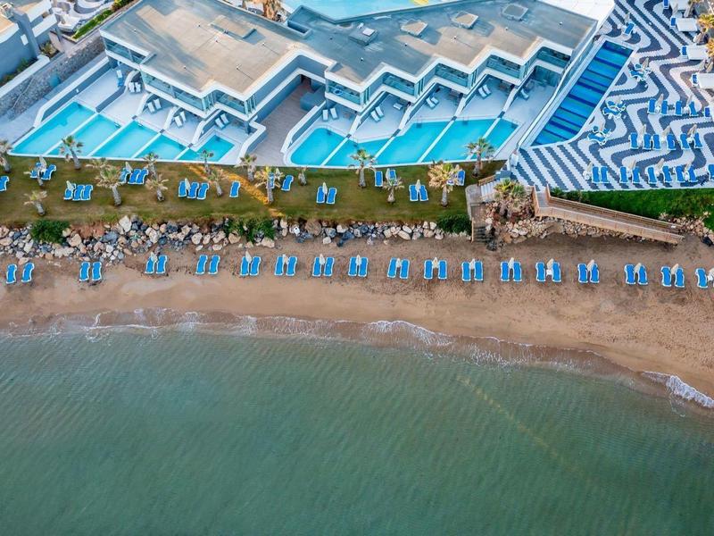 Hotelový komplex se zubatým bazénem a soukromou pláží s modrými slunečníky.