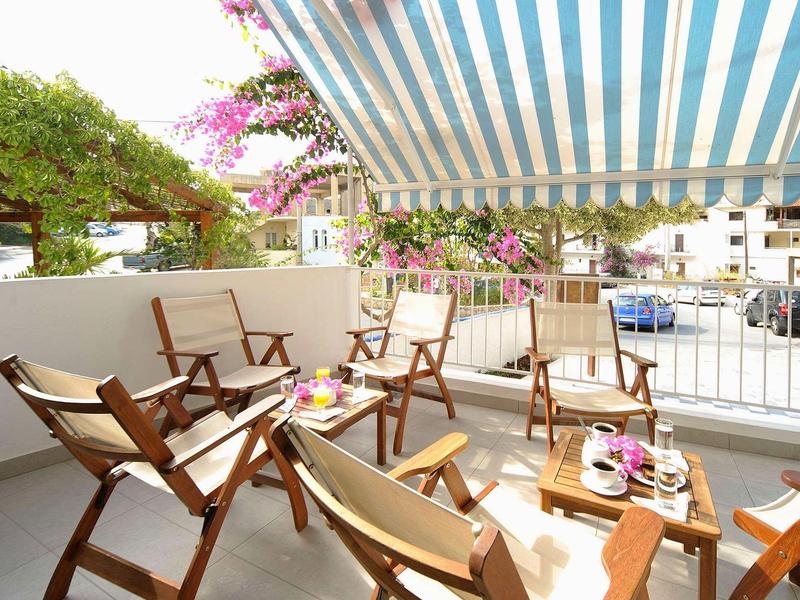 Balkon z drewnianymi krzesłami i stolikiem pod niebiesko-białym pasiastym markizem w słonecznym otoczeniu.