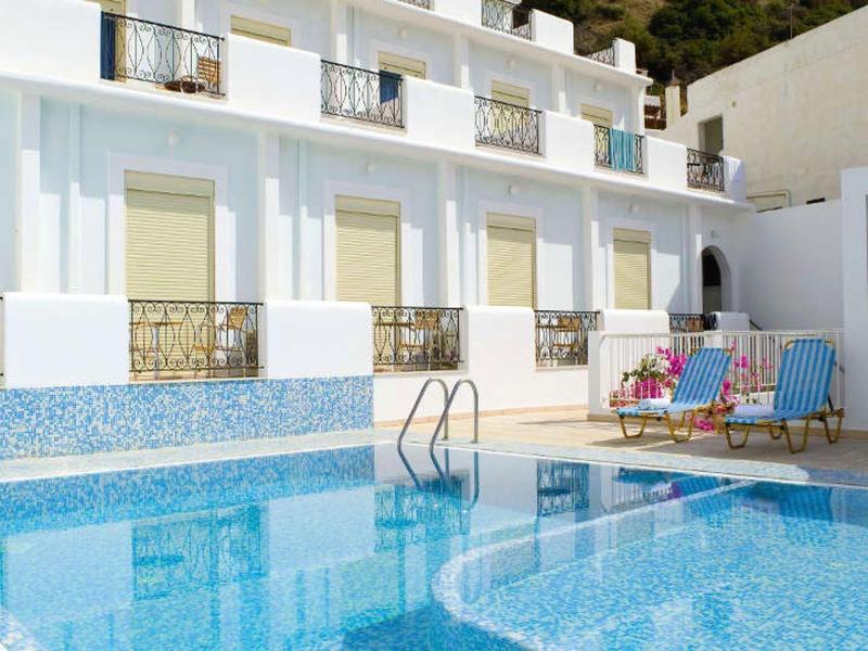 Biały hotel z niebieskim tarasem basenu i leżakami przed zalesionym wzgórzem.