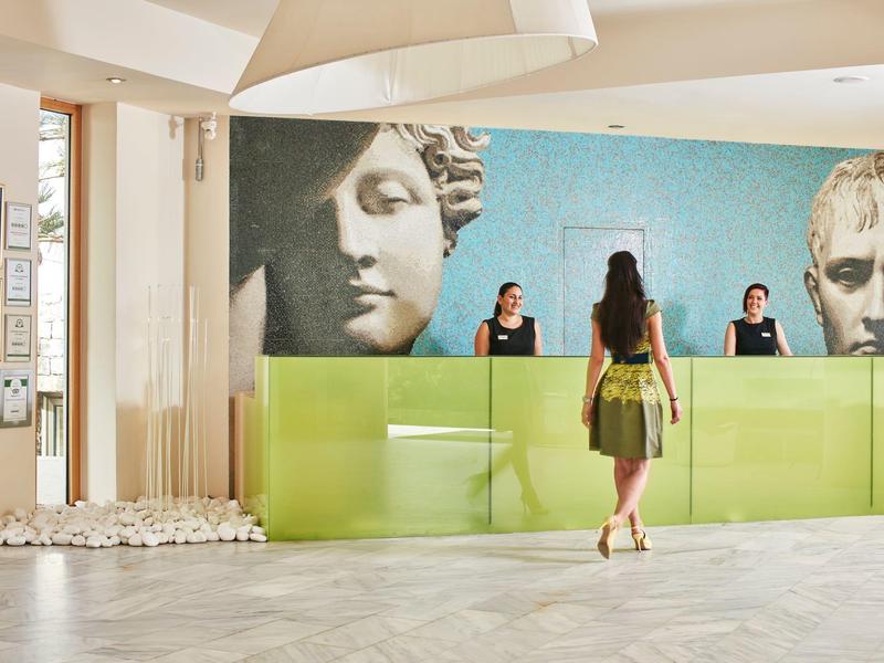 Réception d'hôtel moderne avec comptoir vert, grands portraits artistiques muraux et une femme s'approchant du comptoir.