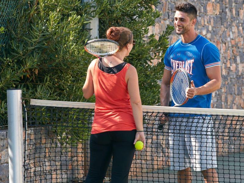 Deux personnes jouent au tennis sur un terrain extérieur avec un mur en pierre en arrière-plan.