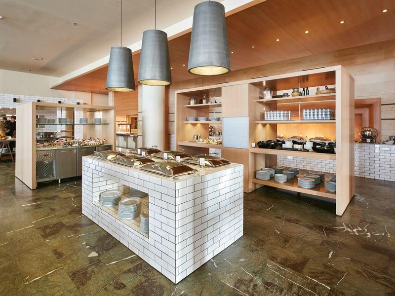 Intérieur de boulangerie moderne avec étagères en bois, îlot en carrelage blanc et lampes suspendues.