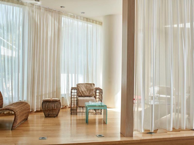 Salon d'hôtel lumineux avec fauteuils en osier, petite table et rideaux blancs transparents près de grandes fenêtres.