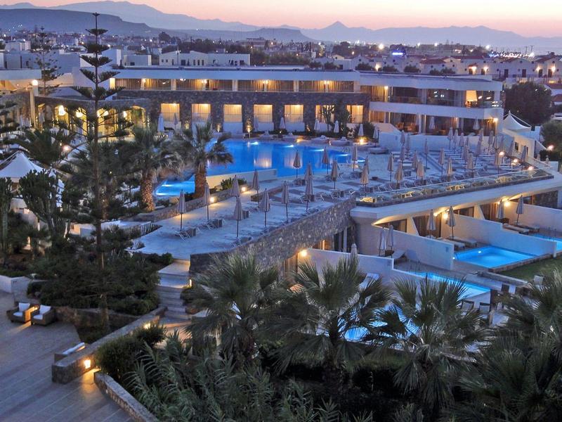 Grand complexe hôtelier au crépuscule avec plusieurs piscines, chaises longues et vue sur la ville.