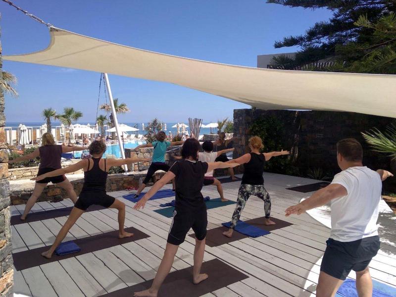 Des personnes font du yoga sur une terrasse en bois près d'une piscine avec des voiles d'ombrage.