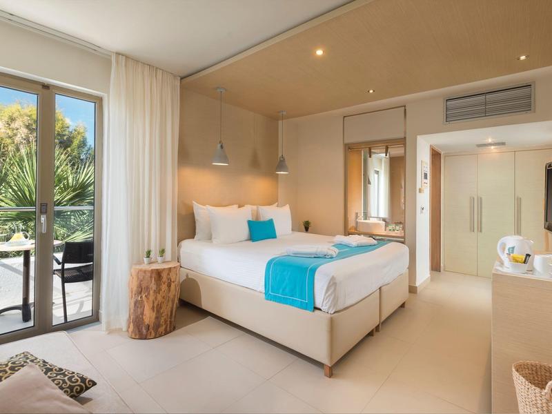 Chambre d'hôtel moderne avec lit blanc et accents turquoise, balcon avec verdure dehors.