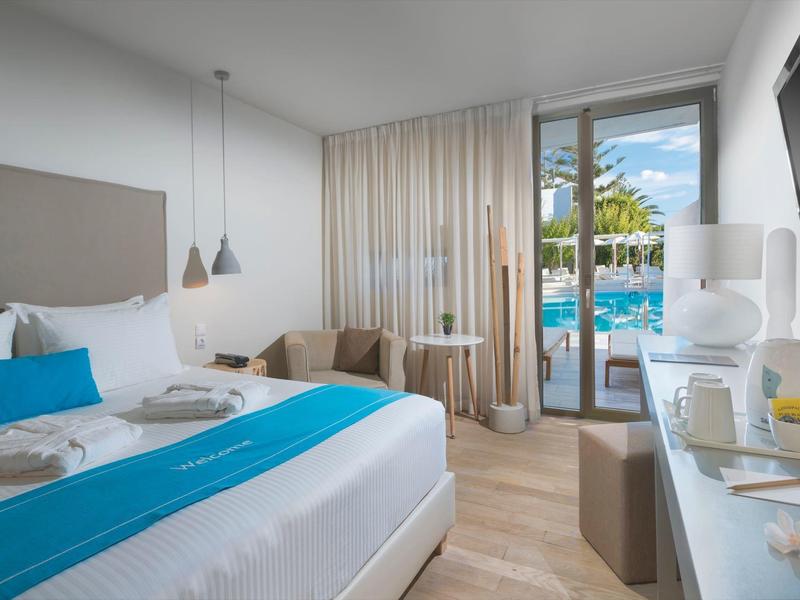 Chambre d'hôtel moderne avec grand lit, accents bleus, coin salon et vue sur la piscine à travers des portes vitrées.