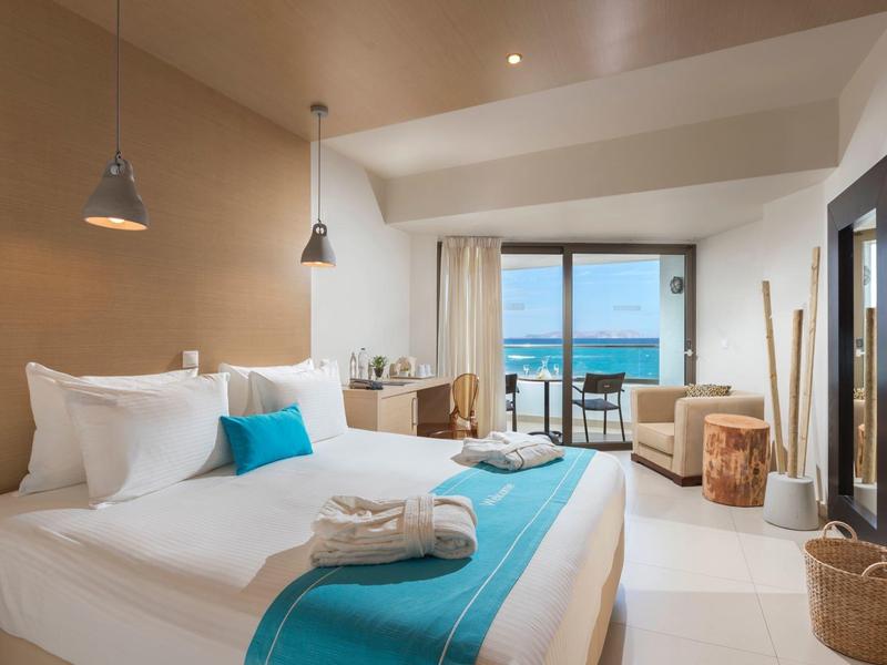 Chambre d'hôtel lumineuse avec lit king-size, accents bleus, balcon et vue sur la mer.