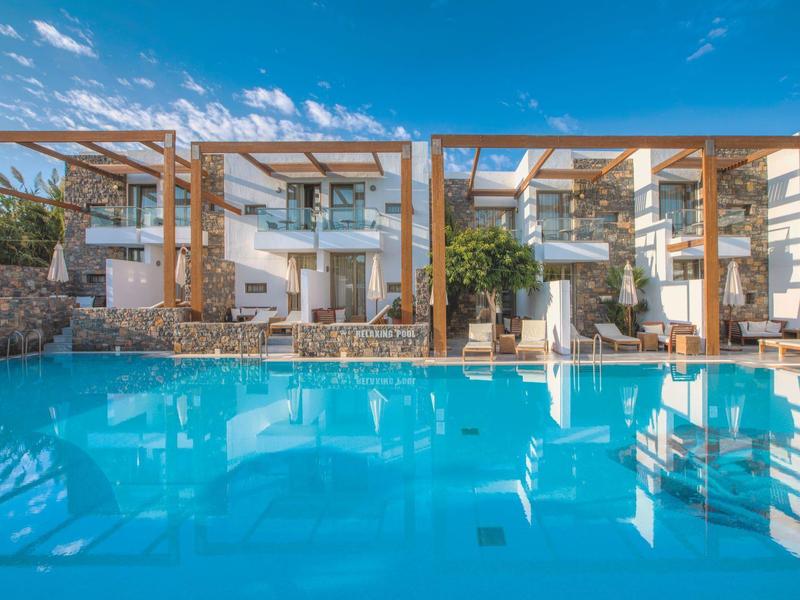Piscine bleue claire avec chambres d'hôtel modernes et pergolas en bois reflétées dans l'eau.