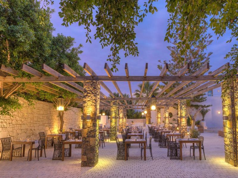 Espace repas extérieur avec pergola en bois, piliers en pierre, tables et chaises sous un feuillage au crépuscule.