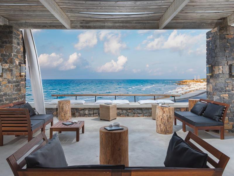 Espace lounge en bord de mer avec meubles en bois et vue sur l'océan entre des piliers en pierre.