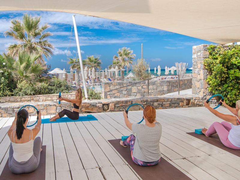 Quatre femmes pratiquent le yoga sur une terrasse en bois avec vue sur la mer et des palmiers en arrière-plan.