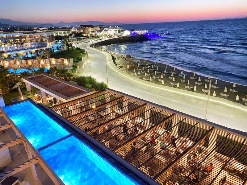 Hôtel en bord de mer au coucher du soleil avec piscine illuminée et coin repas en bord de plage.
