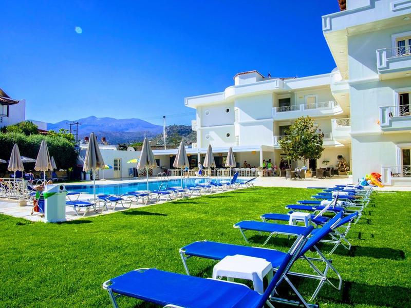 Hotel s venkovním bazénem, lehátky a zeleným trávníkem pod jasnou modrou oblohou.