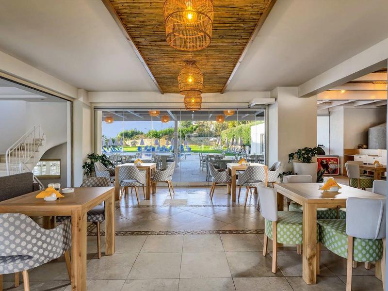 Prosvětlená hotelová restaurace s dřevěnými stoly, vzorovanými židlemi a výhledem na terasu s bazénem.