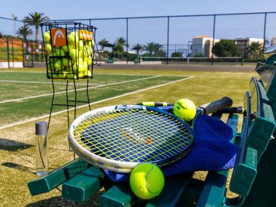 Campo da tennis con racchetta, palline e contenitore per palline su una panchina verde.