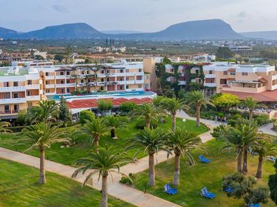 Complesso alberghiero con più edifici, piscina, palme e montagne sullo sfondo sotto un cielo limpido.