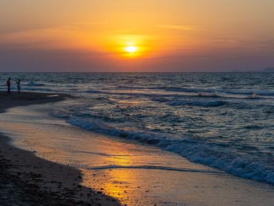 Tramonto sulla spiaggia con onde e due persone sulla riva.