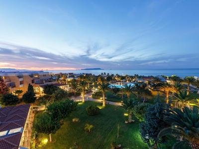 Vista di un resort alberghiero illuminato con giardini verdi e mare sullo sfondo al tramonto.
