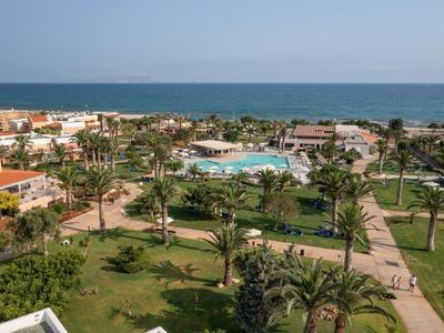Veduta di un resort alberghiero con piscina, palme e mare sullo sfondo in una giornata serena.