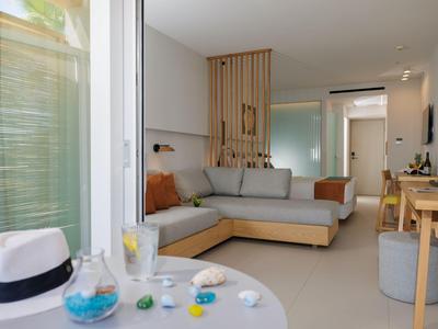 Habitación de hotel luminosa con sofá gris, detalles de madera y balcón con mesa.