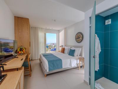 Habitación de hotel luminosa con cama doble, escritorio, TV y baño contiguo con azulejos azules.