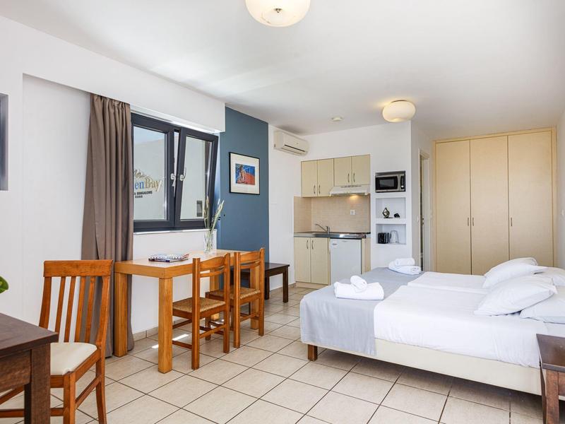Bright hotel room with double bed, dining table with chairs, kitchenette, and large wardrobe.