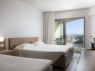 Bright hotel room with three beds, side window, and balcony with outdoor view.