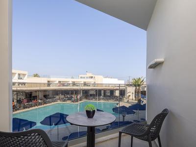 Balcony with table, chairs, and view of hotel pool with blue umbrellas under clear sky.