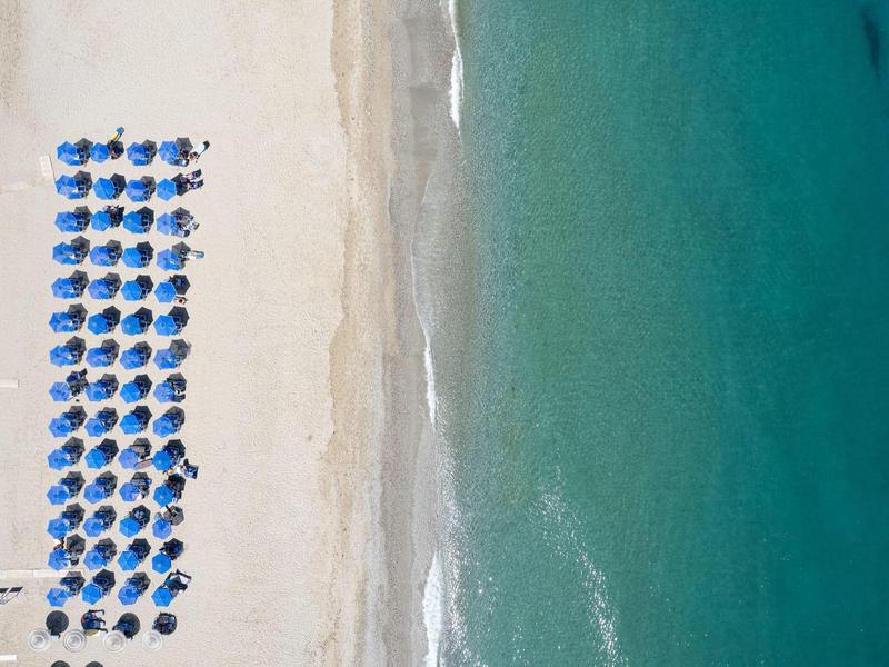 Letecký pohled na pláž s modrými slunečníky a tyrkysovou vodou.