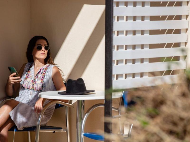 Žena sedí na slunné terase s nápojem, má sluneční brýle.