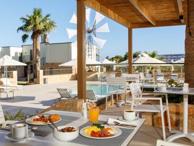 Terrazza con tavolo per la colazione apparecchiato, piscina, palme e mulino a vento sullo sfondo in una giornata di sole.