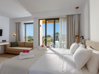 Jasny pokój hotelowy z dużym łóżkiem, częścią wypoczynkową i balkonem z widokiem na palmy.