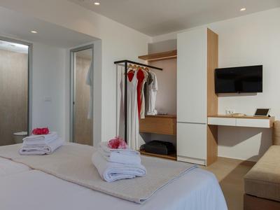 Nowoczesny pokój hotelowy z łóżkiem, telewizorem, otwartą szafą i biurkiem w neutralnych kolorach.