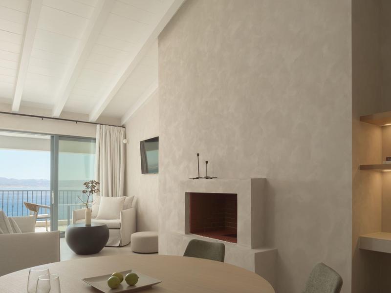 Lichte woonkamer met open haard, eettafel en zeezicht door grote ramen.