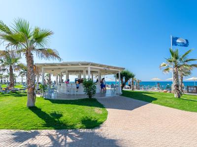 Grünes Parkgelände mit Palmen, Sitzbereich unter Pavillon und Blick auf das blaue Meer und den Himmel.