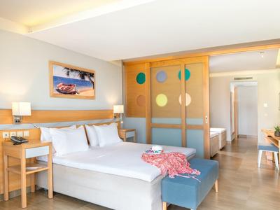 Modernes Hotelzimmer mit Doppelbett, Holzmöbeln, grauem Sitzbank und heller Holztrennwand.