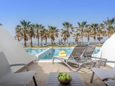 Widok z tarasu z stołami i krzesłami na leżaki, basen i palmy przy morzu.