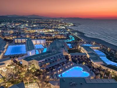 Luftaufnahme eines Küstenresorts bei Sonnenuntergang mit beleuchteten Pools und Meer im Hintergrund.