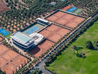 Große Tennisanlage mit mehreren roten Sandplätzen, umgeben von Bäumen und grünem Rasen.