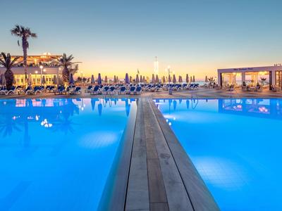 Großer Hotelpool bei Sonnenuntergang mit Holzsteg, Liegestühlen und dekorativer Beleuchtung.