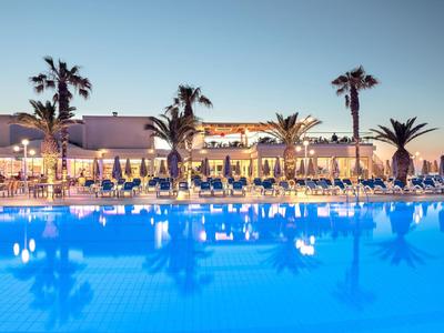 Großer Pool mit blauem Wasser, umgeben von Palmen und einer beleuchteten Terrasse bei Abenddämmerung.